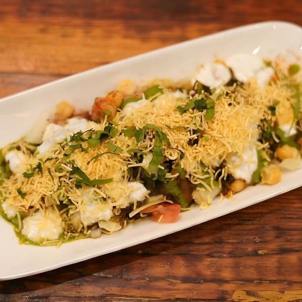 A white rectangular plate filled with Indian chaat, topped with yogurt, green chutney, and sev.