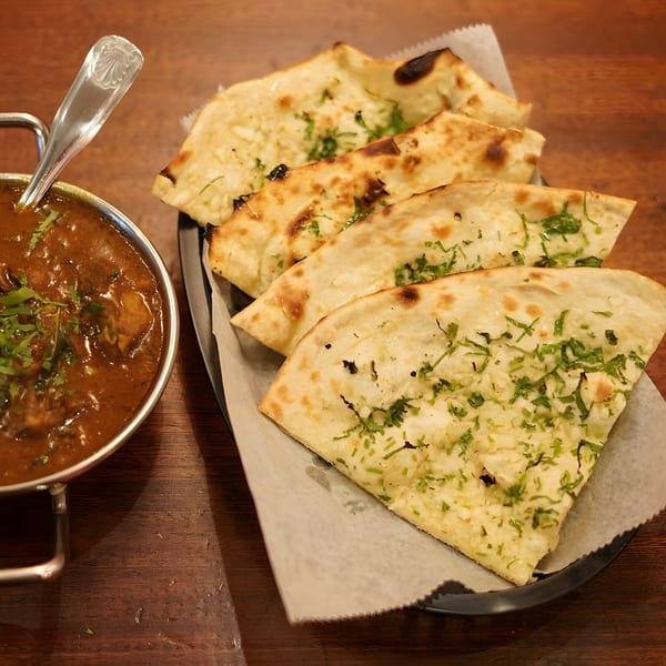 Indian curry in a metal bowl with a spoon, alongside a basket of garlic naan bread.